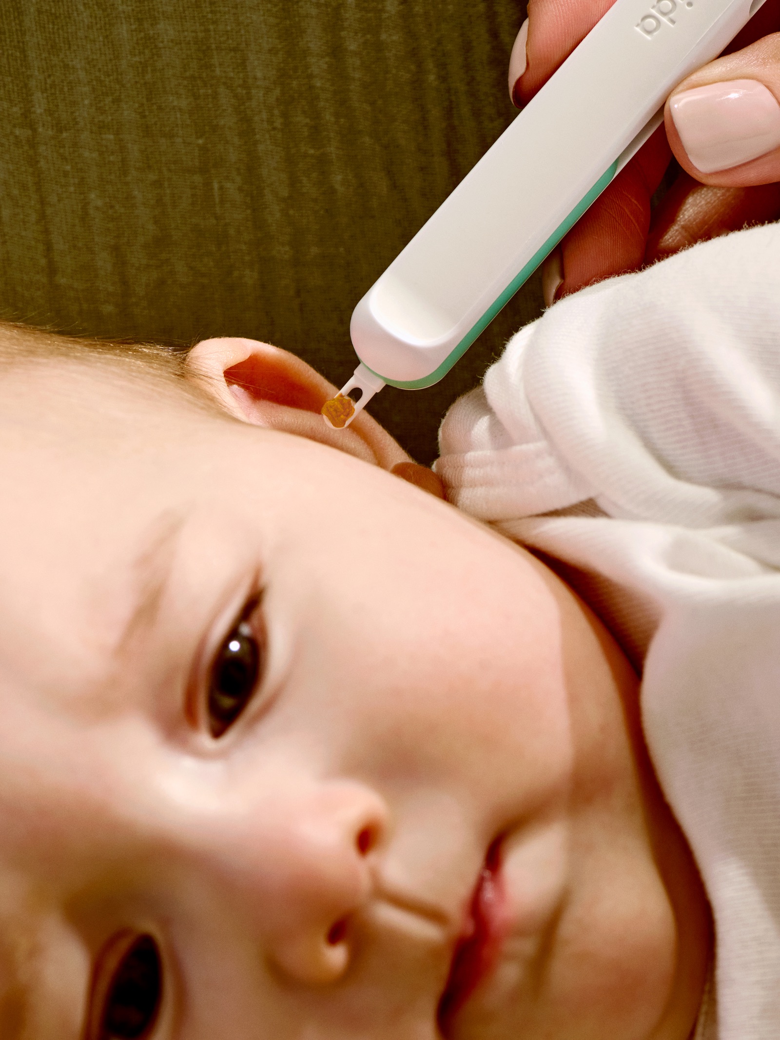 A parent gently cleaning the outer ear of a baby with a Frida Baby grooming tool.