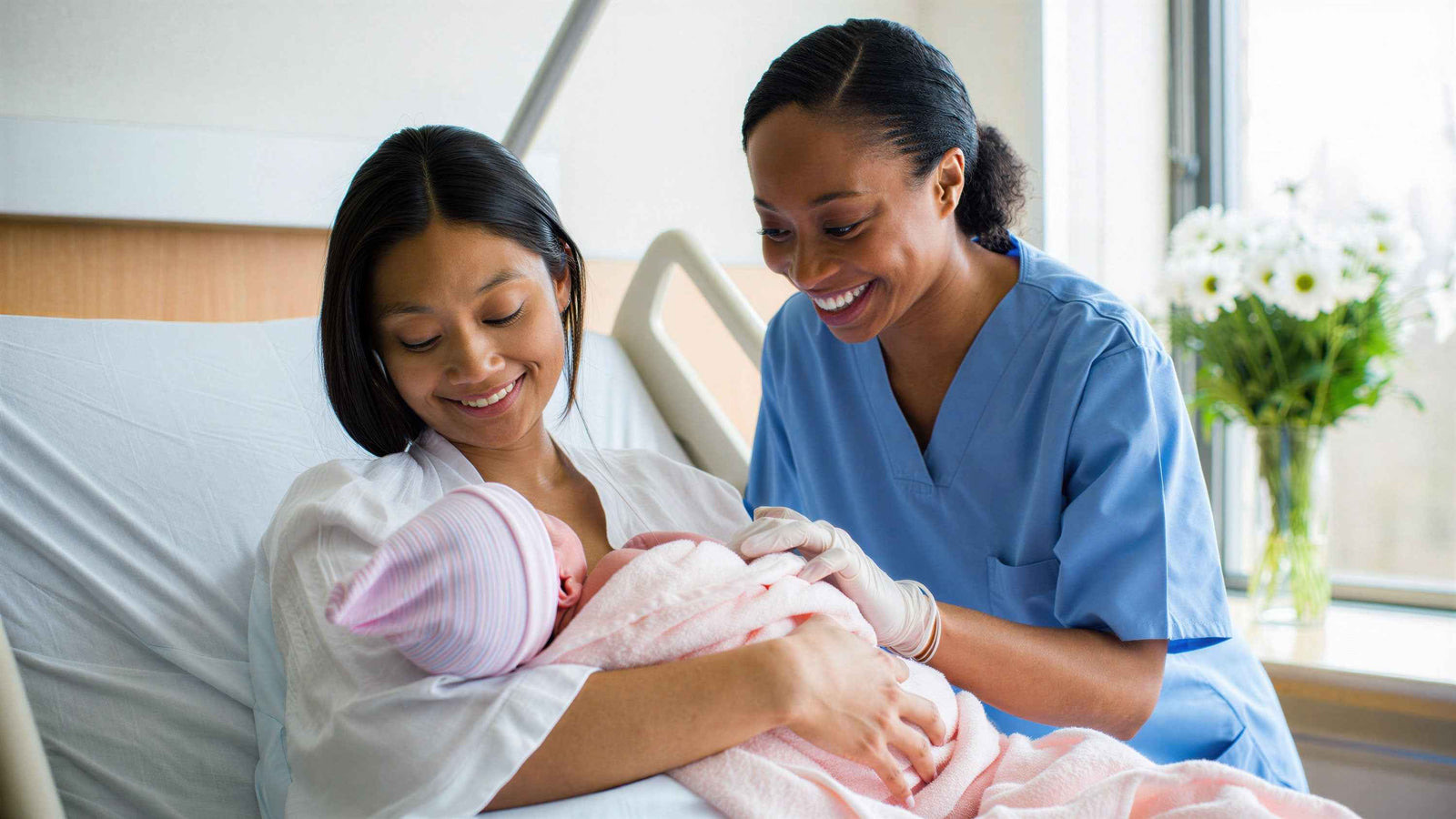 A new mum holding her newborn in a hospital bed while a smiling midwife sits beside her.