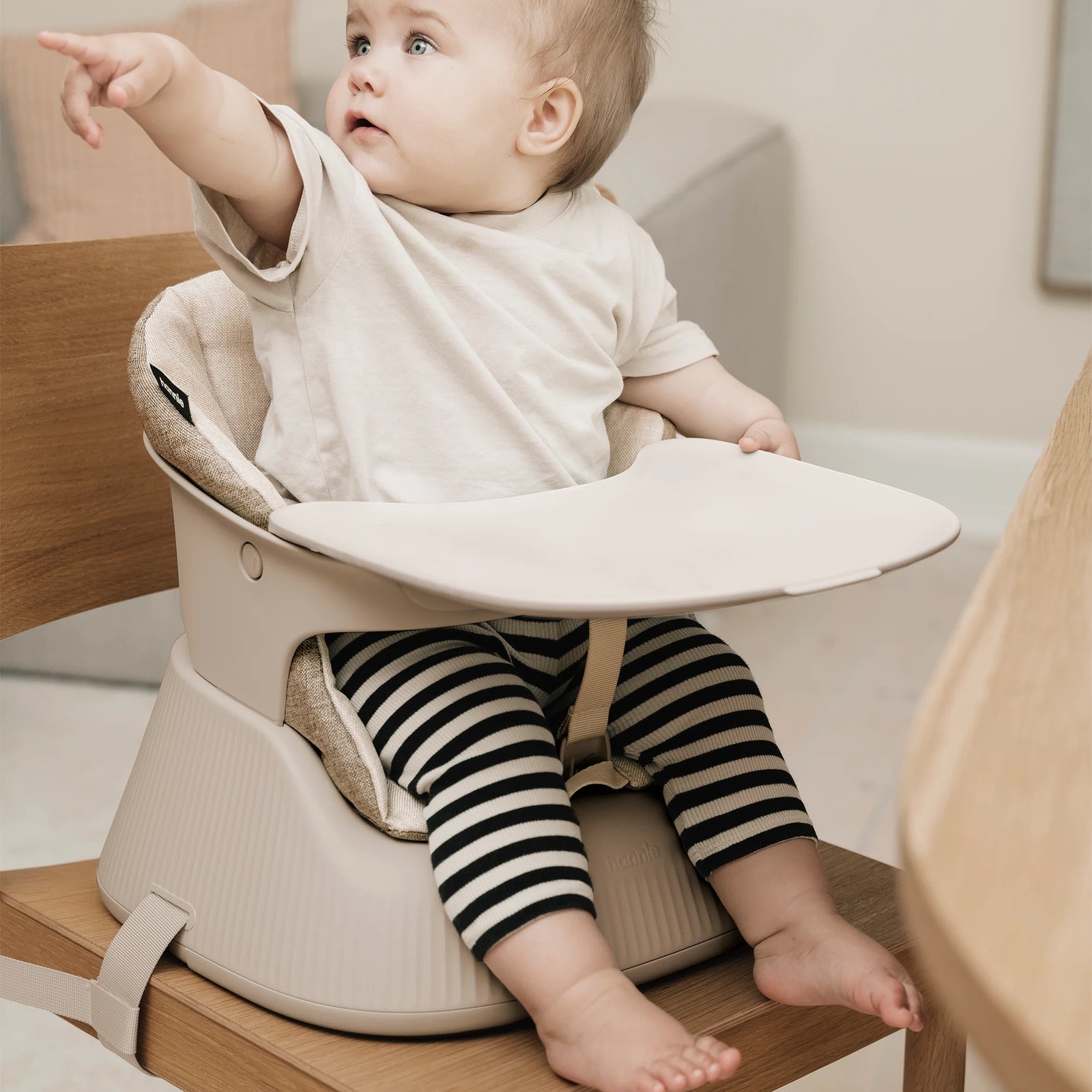 Baby secured with the harness in a White Sand Hannie with the tray on, beside a timber table