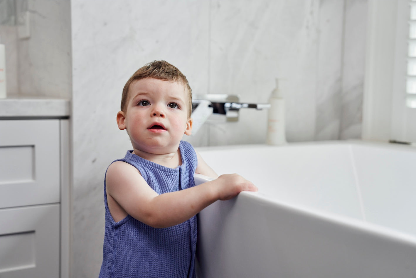 A toddler standing beside a bath in a bright white bathroom.
