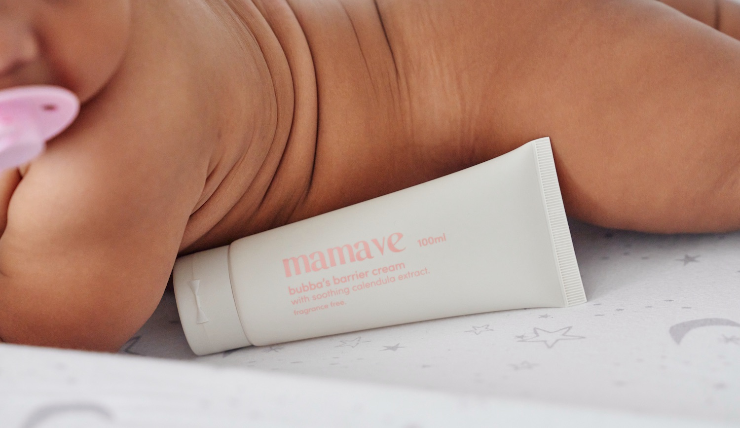 A baby lying on a star print sheet beside a tube of Mamave Bubba's Barrier Cream.