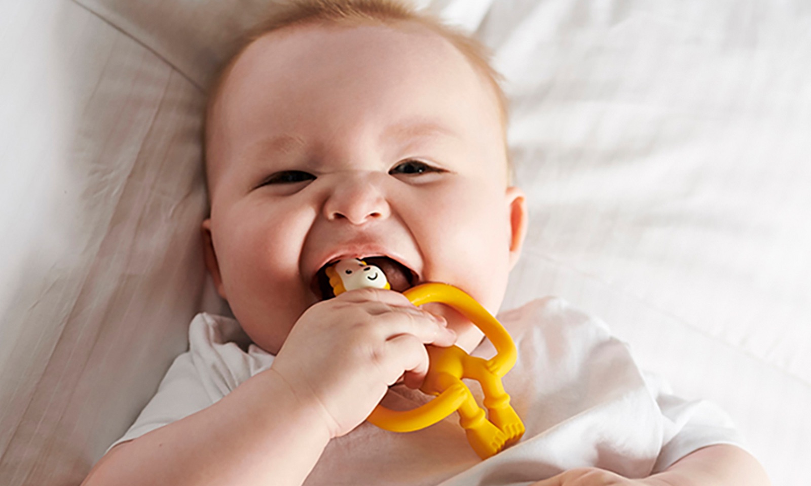 Baby lying on a bed chewing a yellow Matchstick Monkey teether.