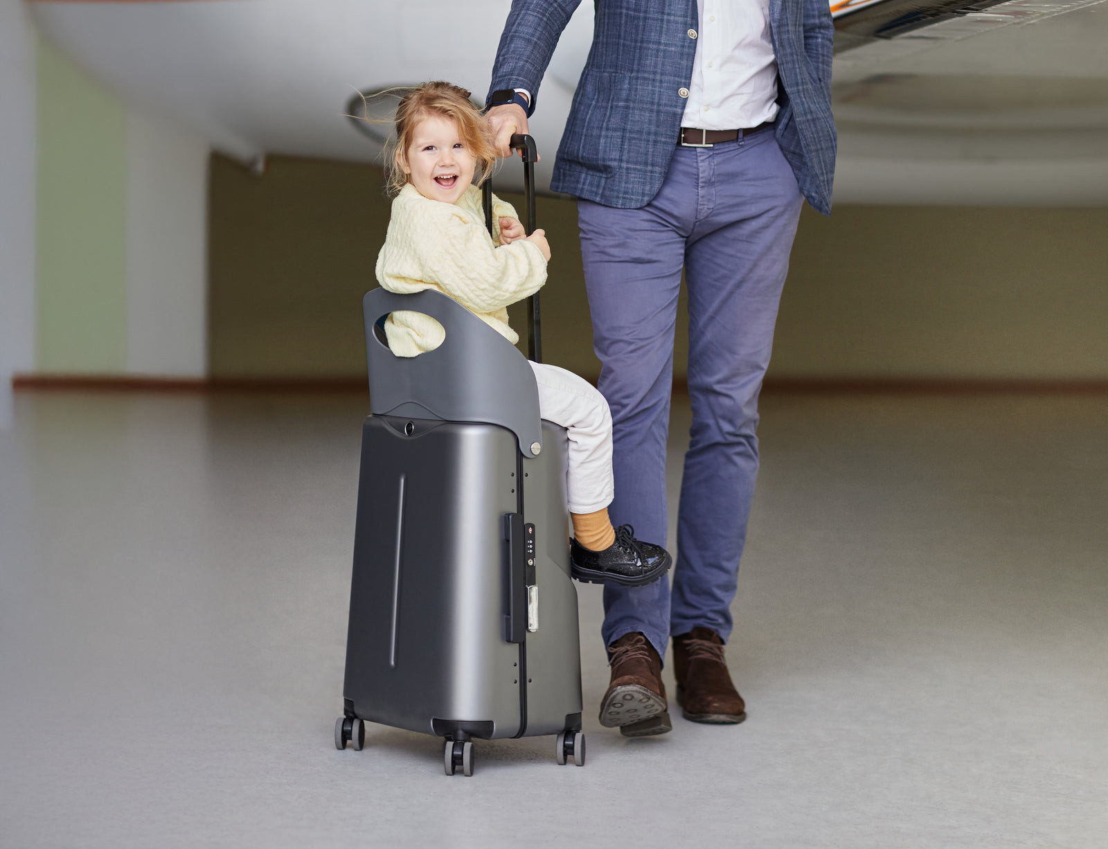 Toddler riding on a grey MiaMily suitcase pulled by a parent indoors