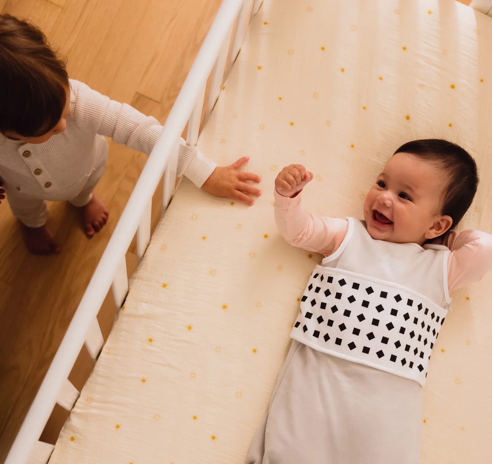 Smiling baby lying in a cot wearing a Nanit Breathing Wear Band, with an older sibling reaching through the rails.