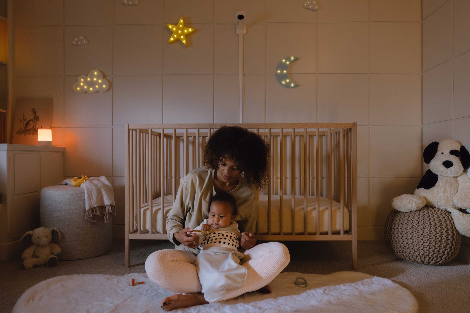 Mother sitting on the nursery floor with her baby in front of a cot, with a Nanit camera above the cot in soft evening light.