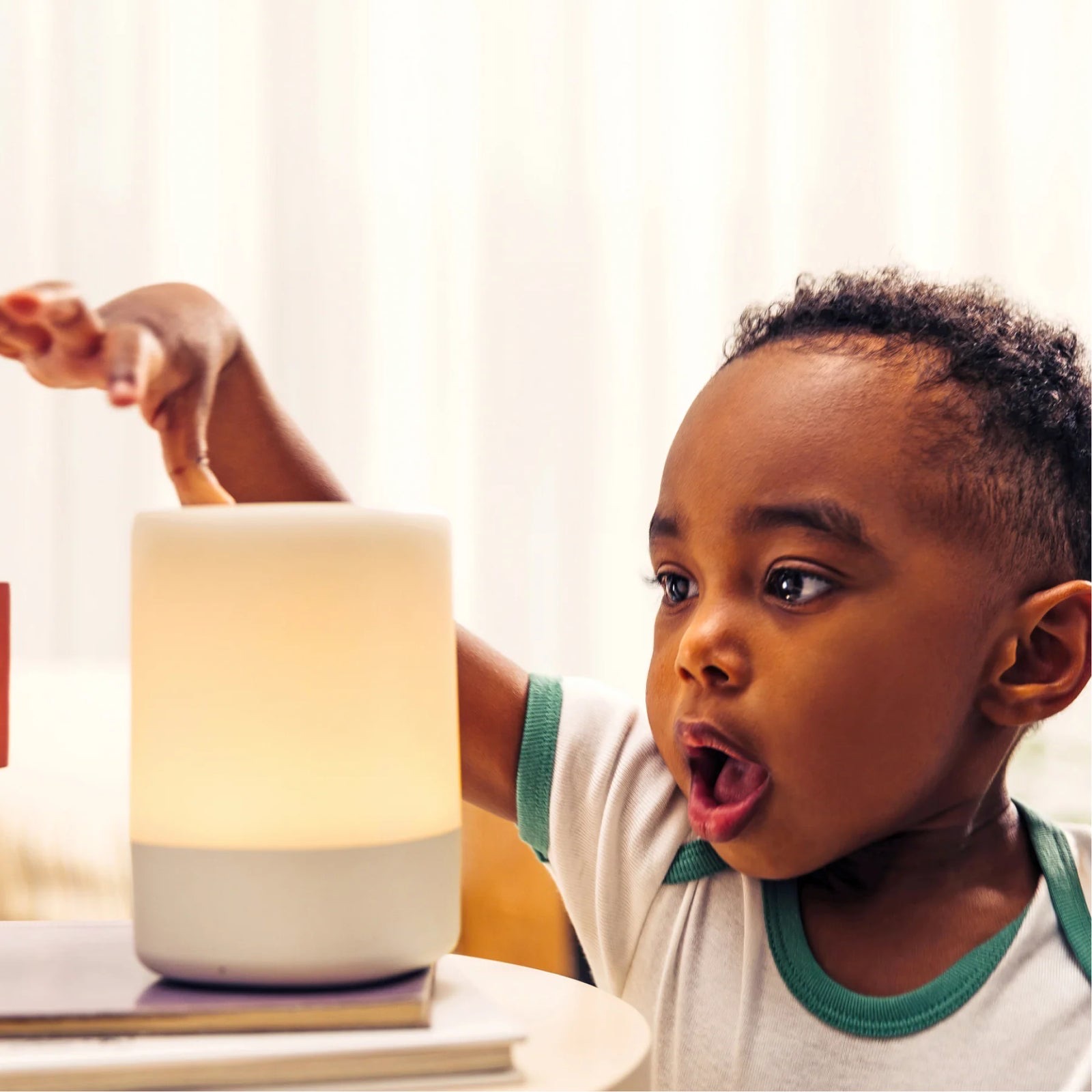 Toddler reaching up to touch a glowing Nanit Sound + Light Machine on a small table.
