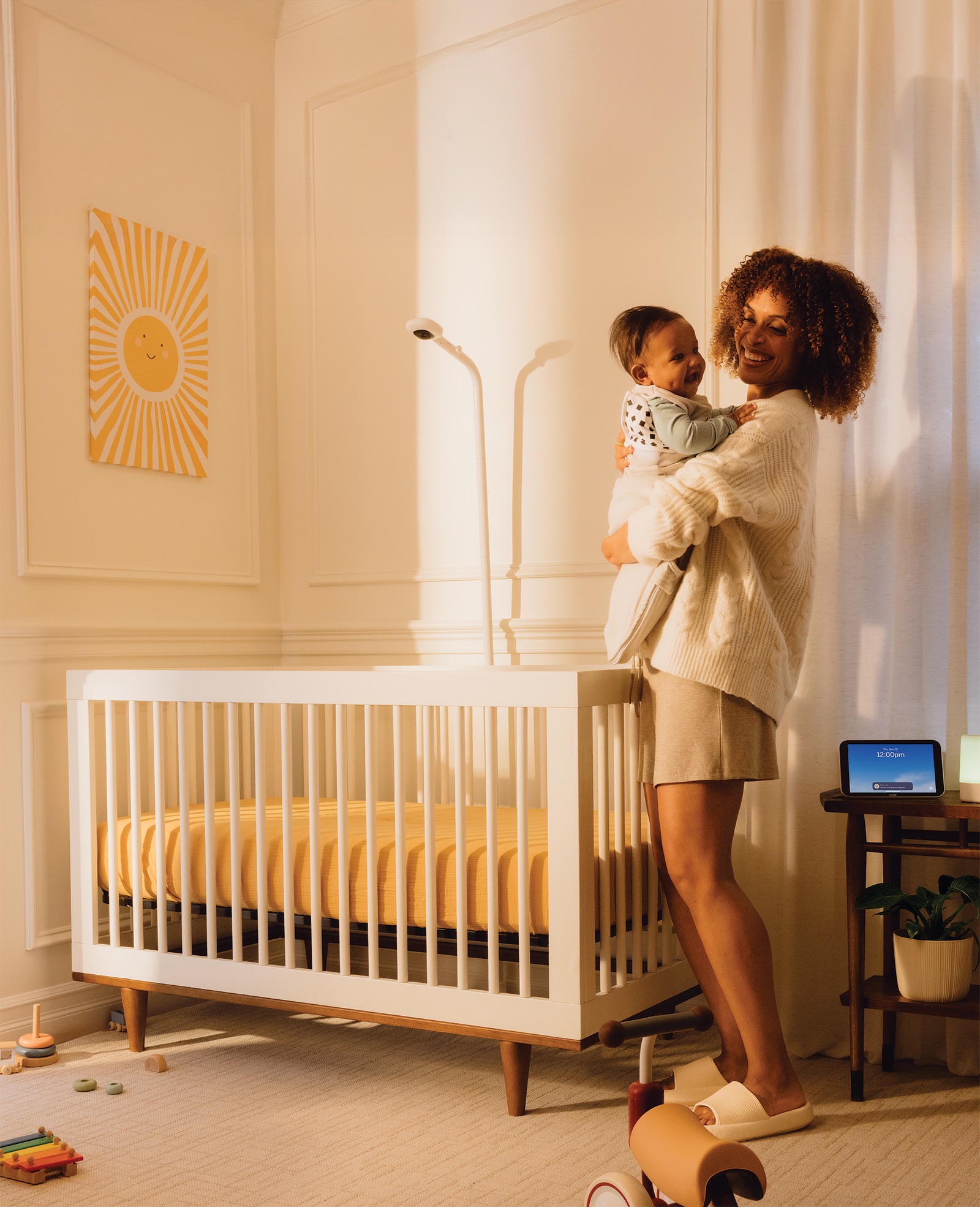 Mother holding her smiling baby beside a cot with a Nanit camera on a Floor Stand above the mattress.