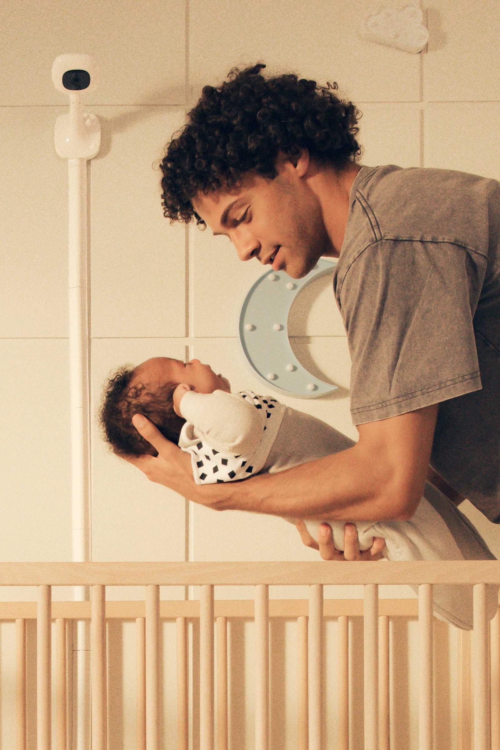 Father holding his newborn above the cot, beneath a Nanit camera mounted on the nursery wall.