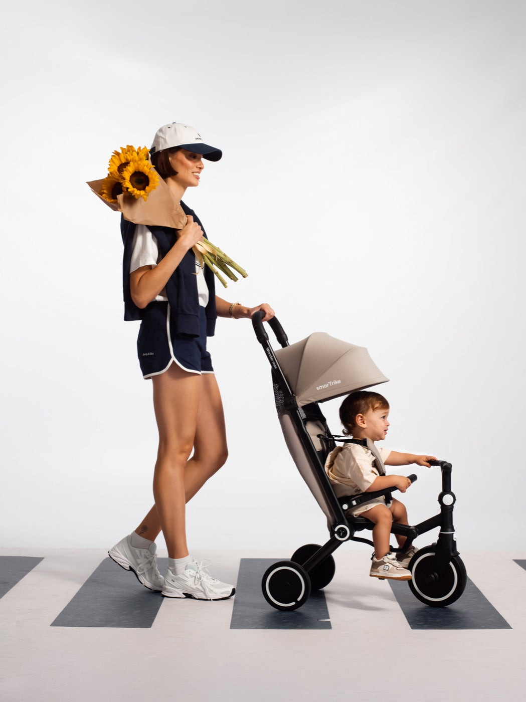 A parent carrying sunflowers pushes a toddler in a Stone Beige smarTrike Wonder max across a pedestrian crossing.
