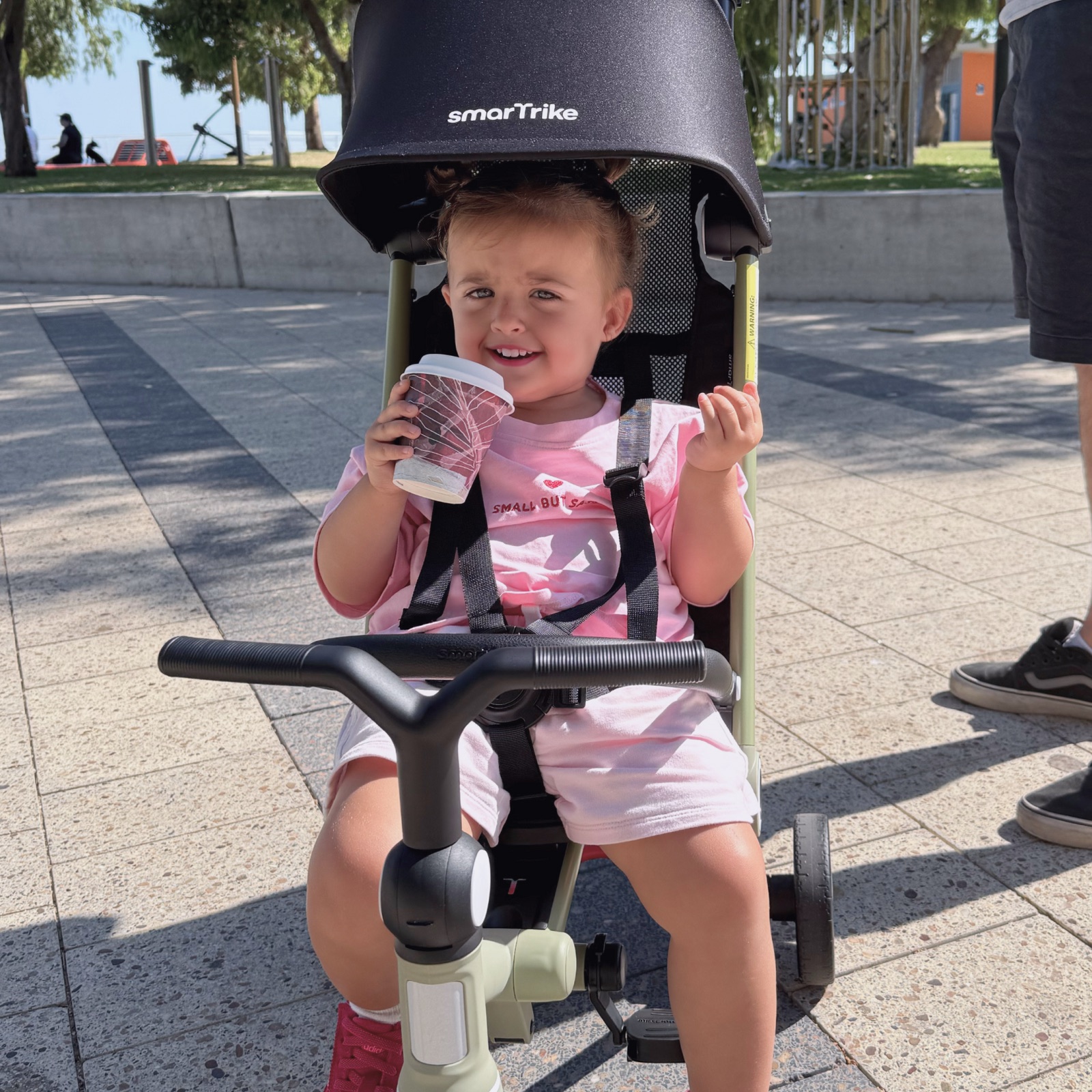 A smiling toddler sits under the canopy of a smarTrike stroller trike on a sunny paved walkway.