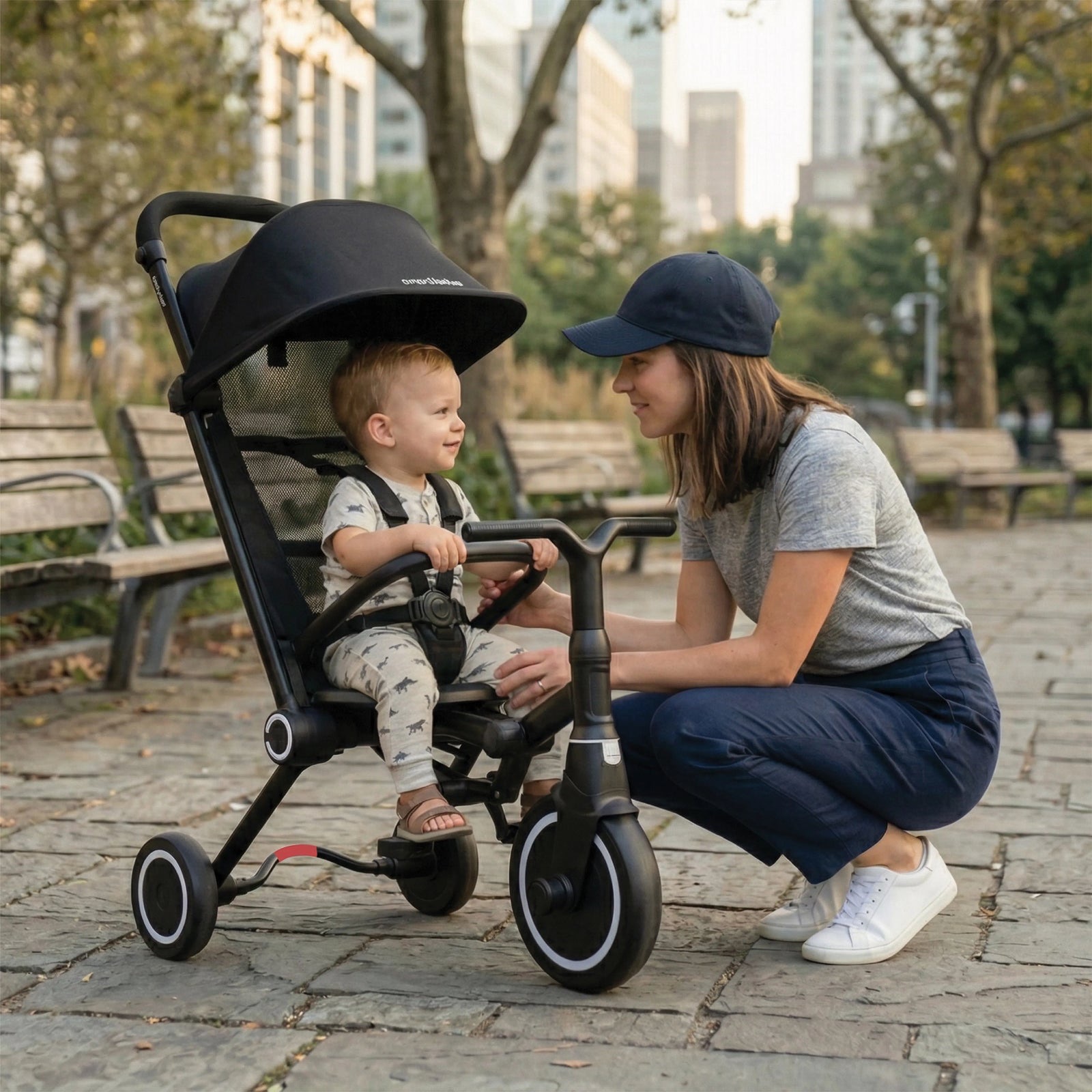 A parent crouches beside a smiling toddler sitting in a black smarTrike Wind range trike in a city park.