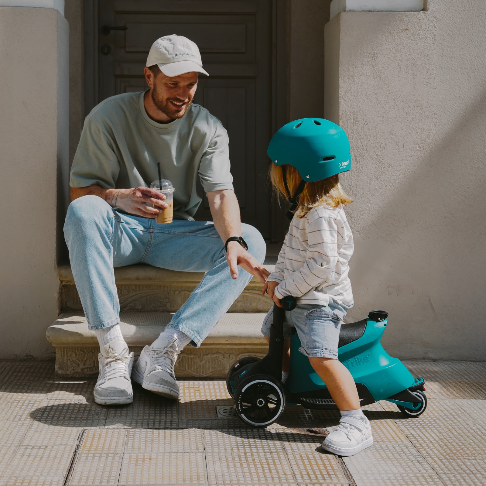 A father sits on a doorstep while a toddler in a helmet sits on a teal smarTrike Xtend Ride-on.