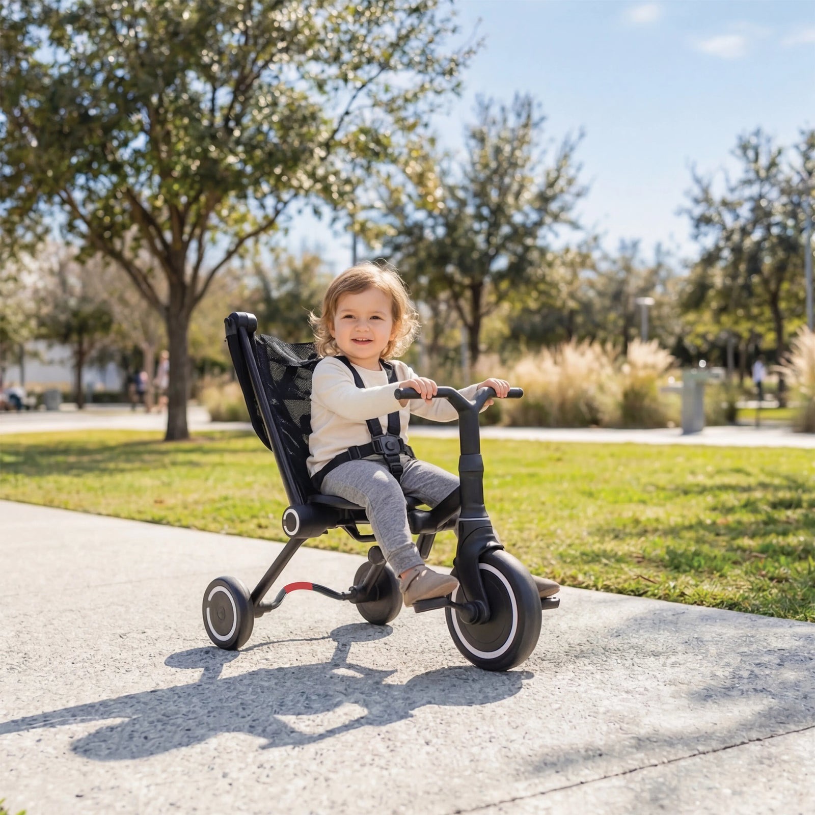 A smiling toddler sits in a black smarTrike Wind range trike on a sunny park path.