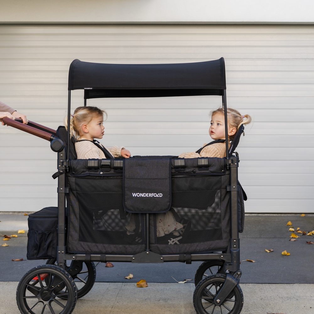 Two toddlers sitting face to face in a black WonderFold stroller wagon