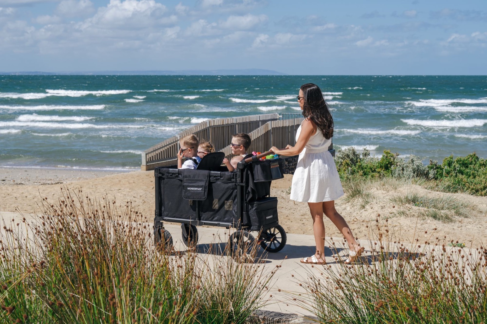 Parent pushing a WonderFold stroller wagon full of children along a beach path