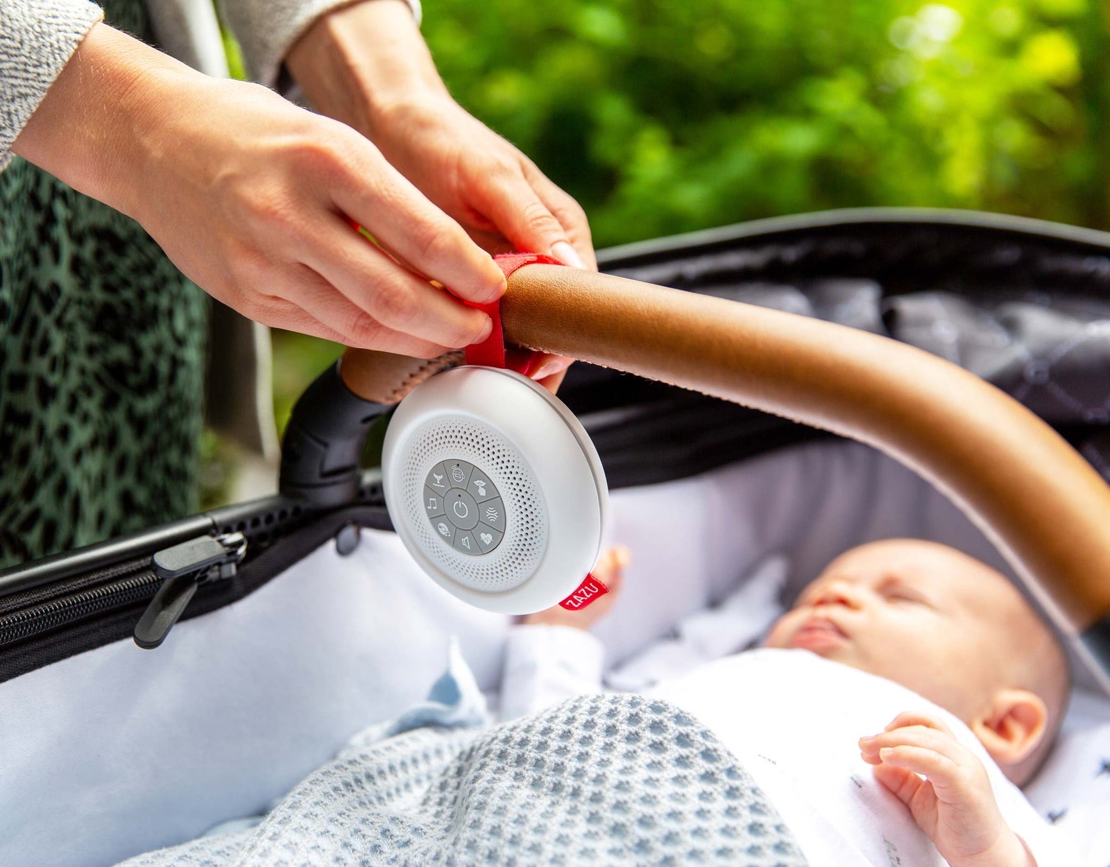 A parent fastening the Suzy soother to a pram handlebar above a sleeping baby.