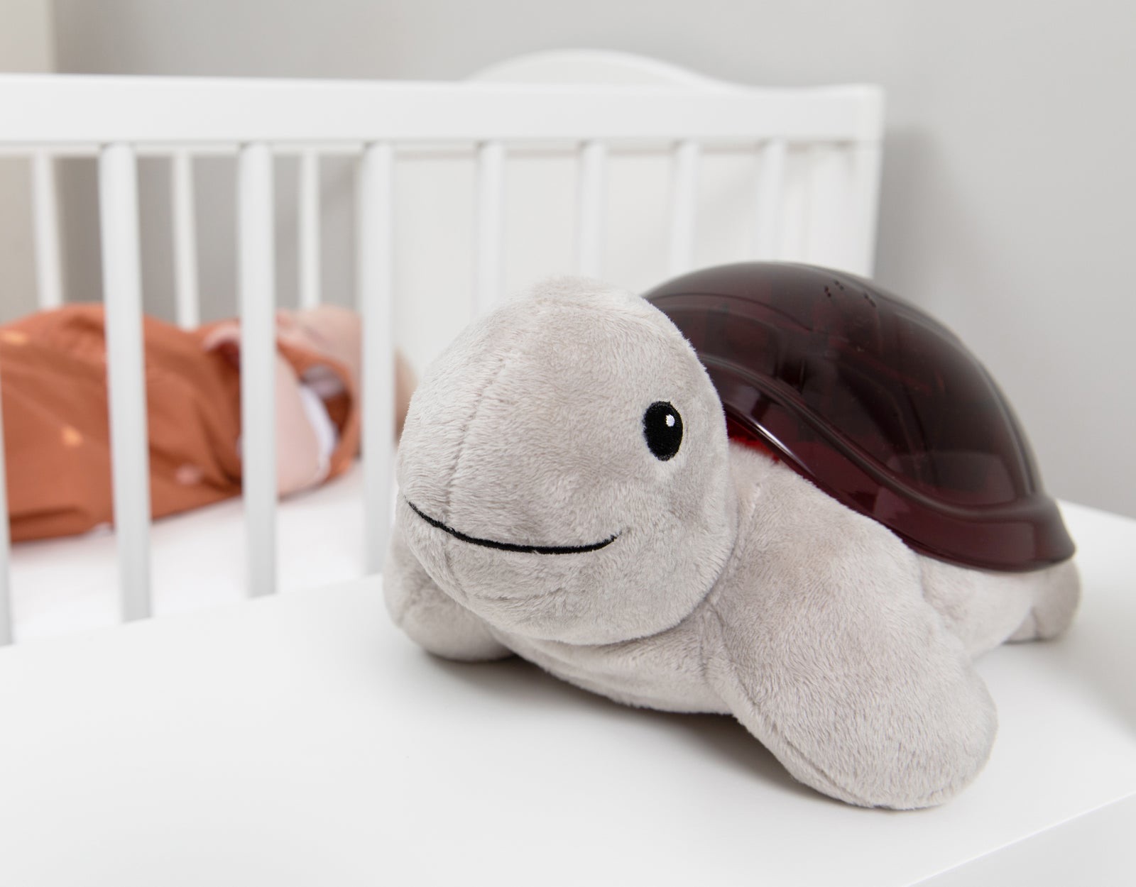 Tim the Turtle sunset projector on a white shelf beside a cot with a sleeping baby.