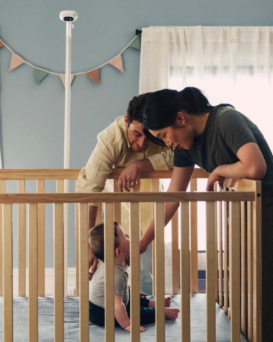 Parents settling a toddler in a cot under a Nanit camera