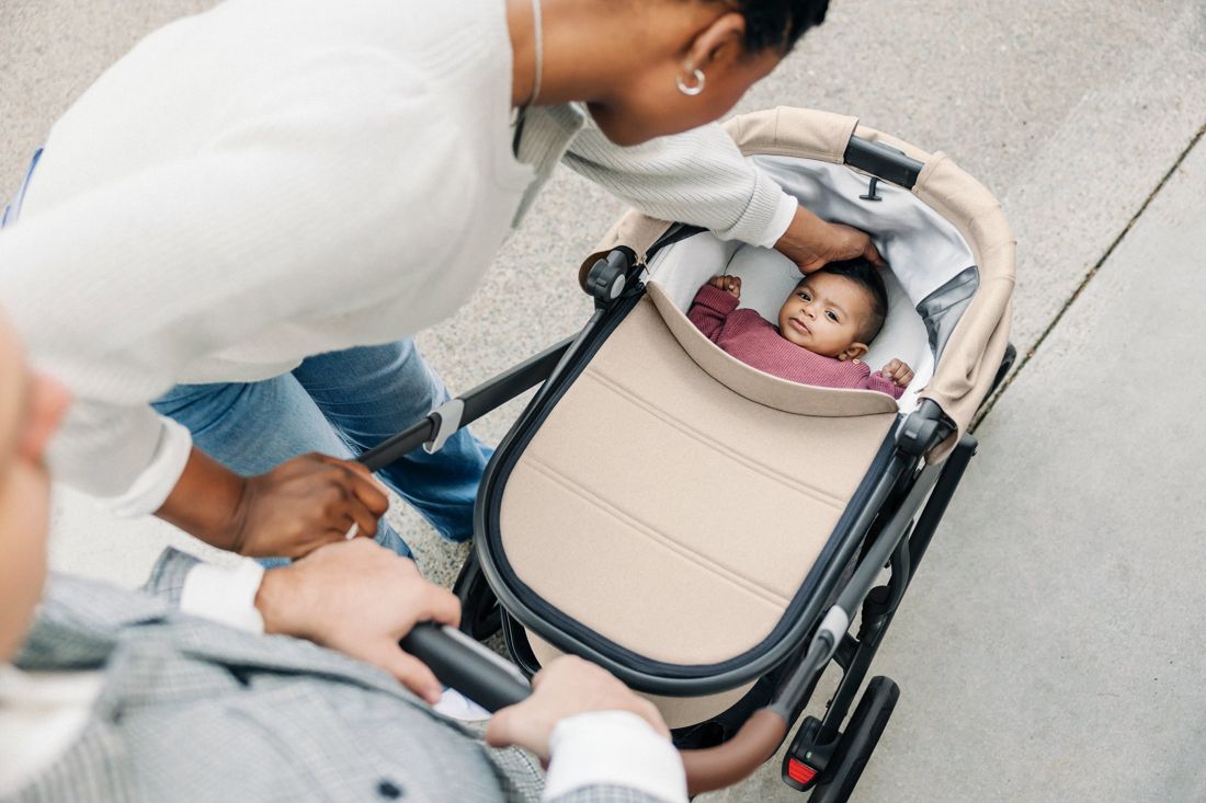 Overhead view of parents pushing a sand-coloured bassinet on a pram as mum strokes the baby's head on a concrete footpath