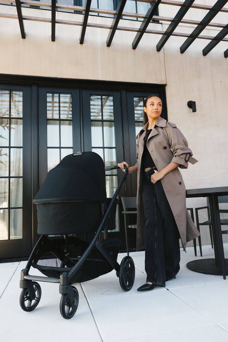 Mum in a trench coat stands with a black Cruz V3 fitted with the bassinet on a terrace beneath a pergola