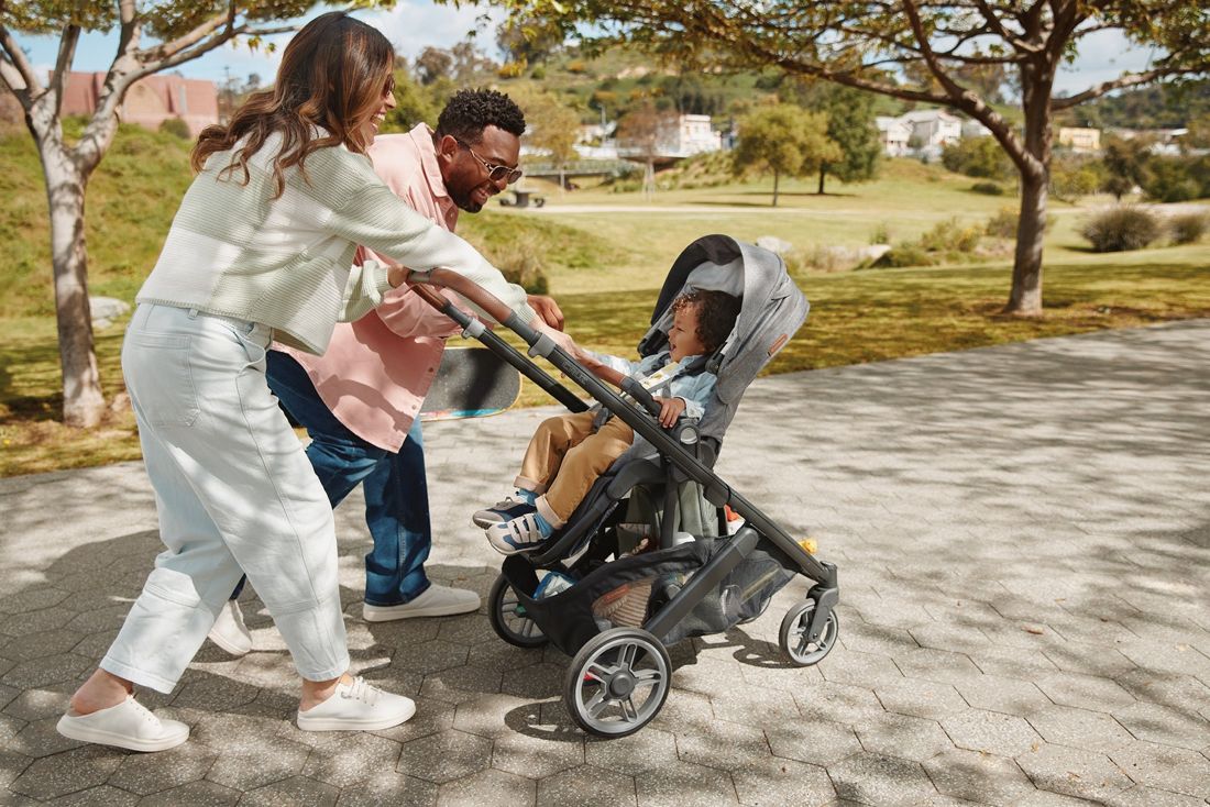 A mum and dad push a grey Cruz V3 with a toddler in the seat along a park path.