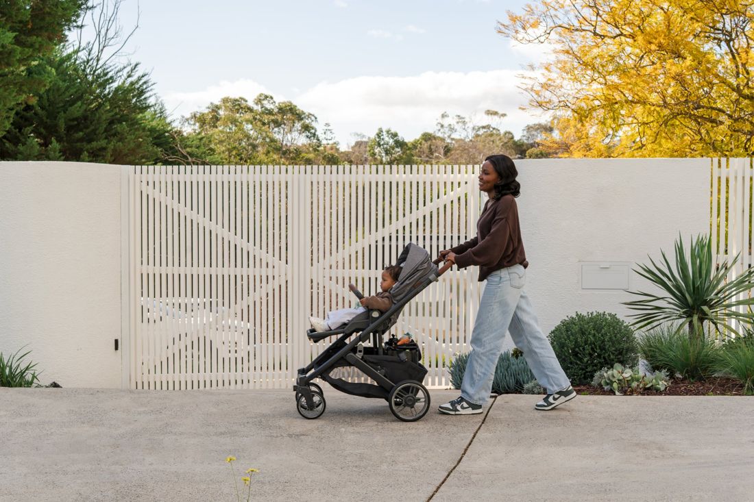 Mum in a brown jumper and jeans walks a Cruz V3 with a toddler past a white slatted fence under blue sky, Australian gum trees behind