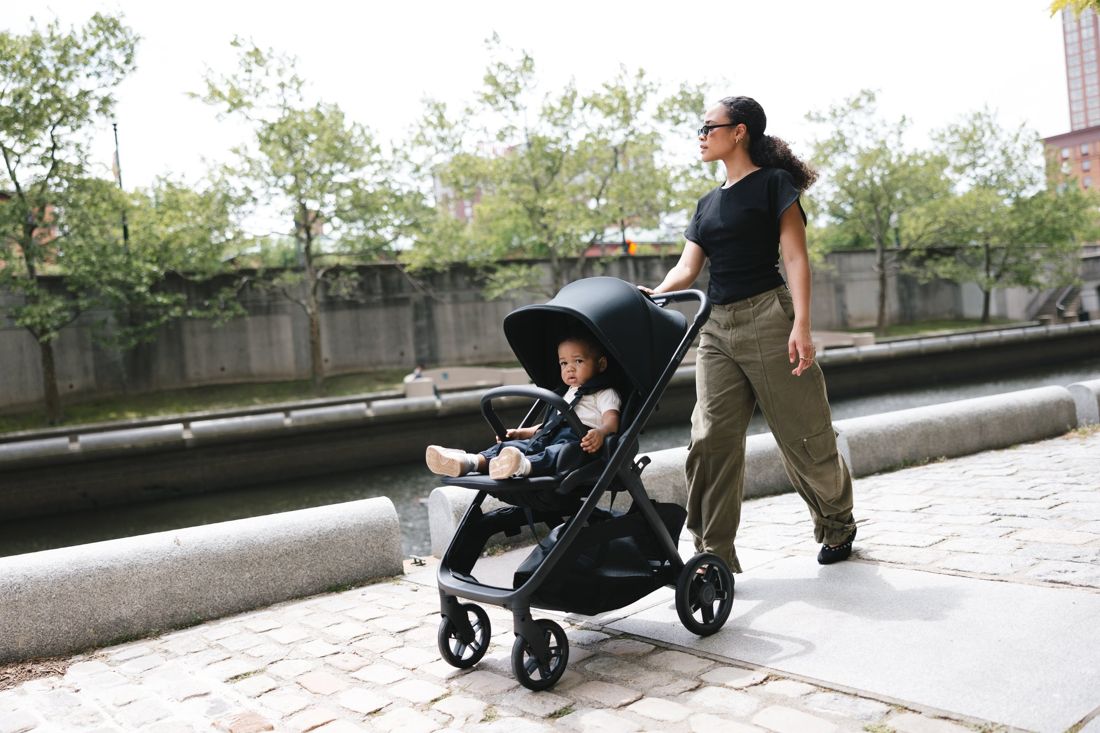 A mum in sunglasses pushes a black Kona with a toddler facing forward along a cobbled riverside path.