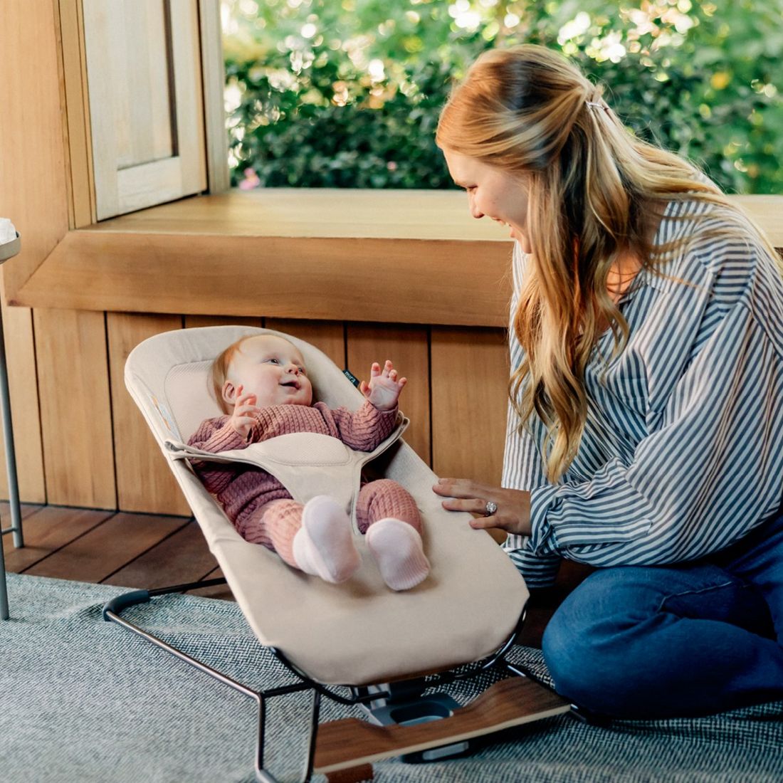 A mum sits on the floor beside a smiling baby in a sand-coloured Mira baby bouncer