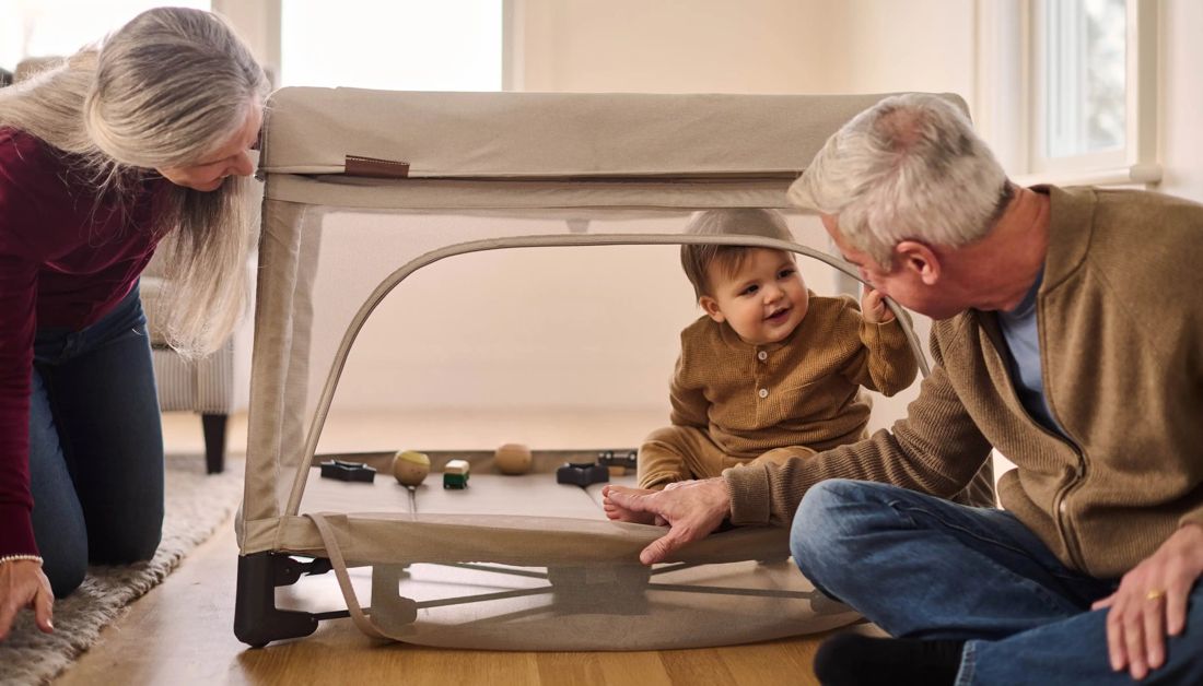 Grandparents kneel on the floor either side of a Remi Portacot as a smiling toddler peeks through the open mesh door