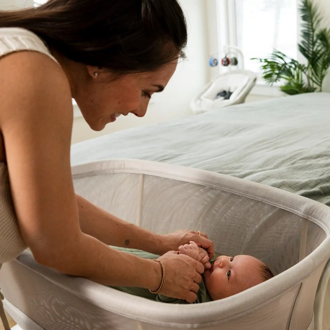 A mum leans into a Soma bassinet, holding the hands of a swaddled newborn who looks up at her