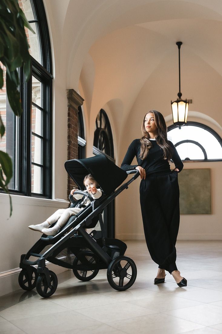 A mum leans on a black Vista V3 with a smiling toddler in the seat in a bright arched hallway.