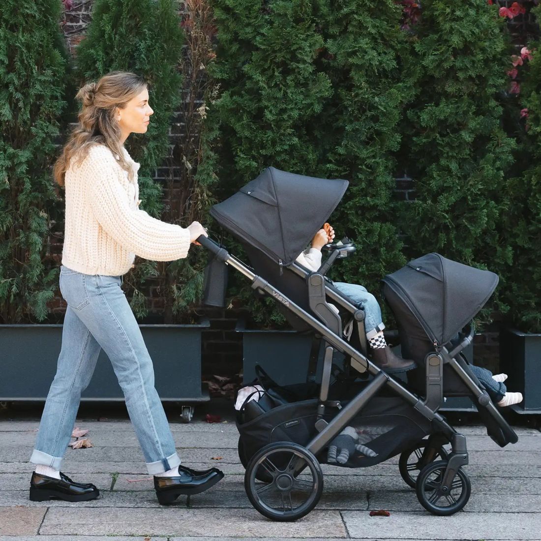 Side view of a mum walking a charcoal Vista V3 with two children in the toddler seat and RumbleSeat along a hedge-lined footpath
