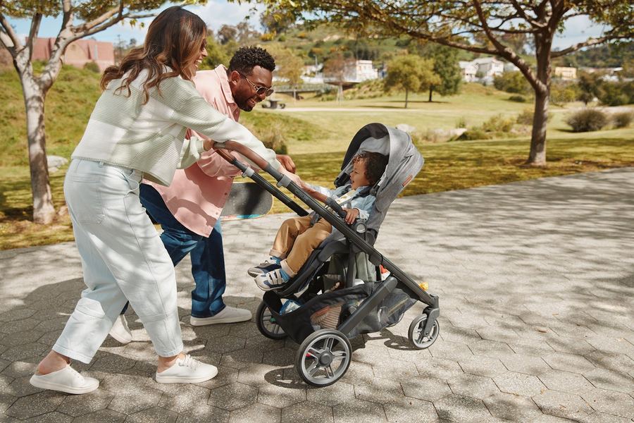 Laughing mum and dad push a grey Cruz V3 with a happy toddler in the seat on a sunny park path