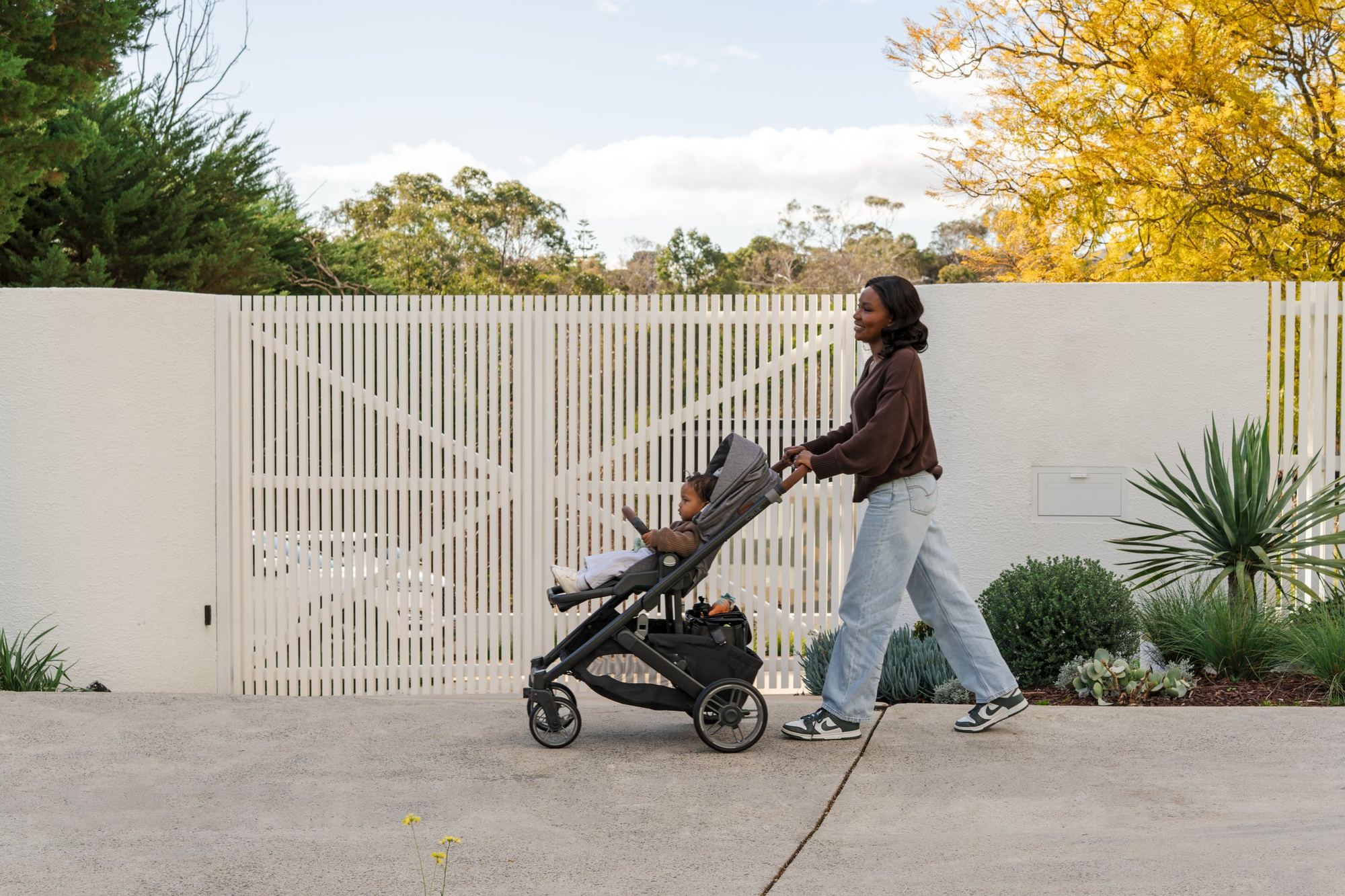 Mum in a brown jumper and jeans walks a Cruz V3 with a toddler past a white slatted fence under blue sky, Australian gum trees behind