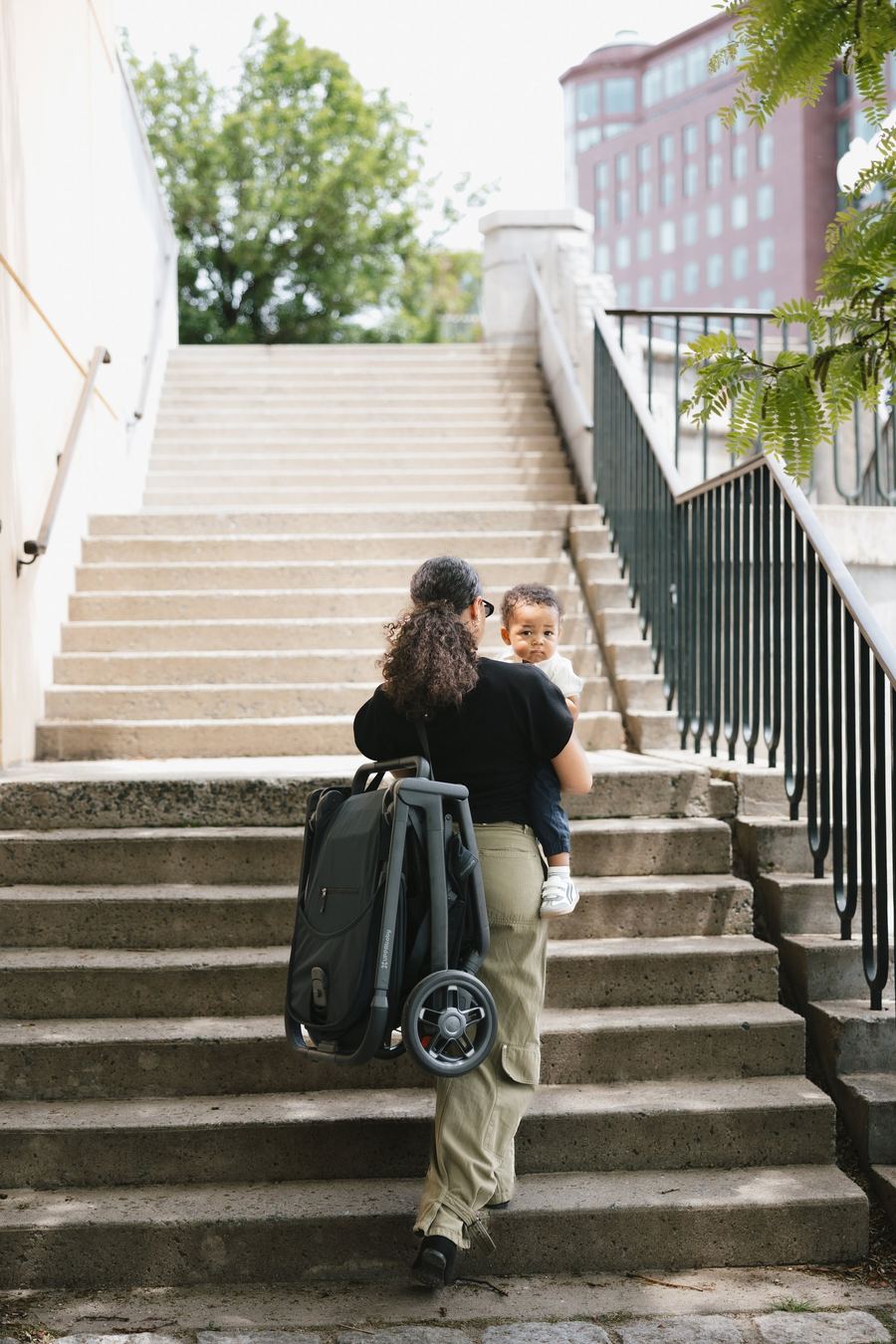Seen from behind, a mum climbs outdoor concrete stairs carrying a toddler on her hip and the folded Kona over her shoulder