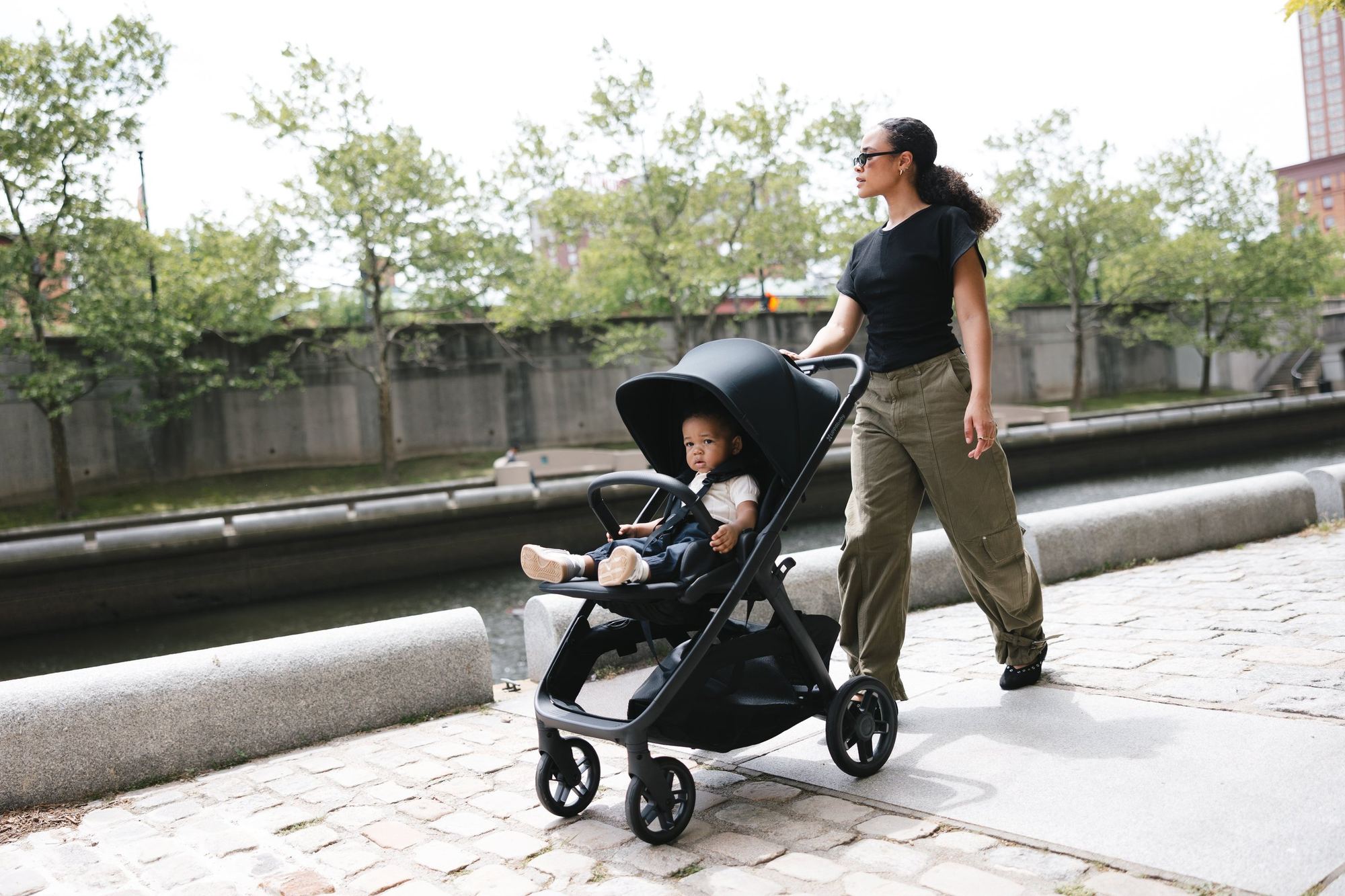 Mum in sunglasses pushes a black Kona with a toddler seated forward-facing along a cobbled riverside path lined with trees