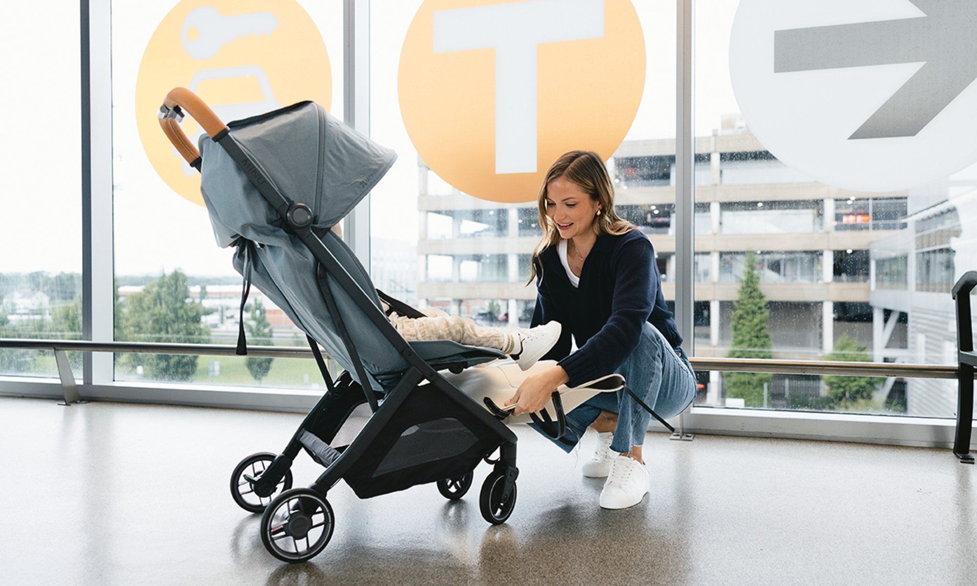 Mum crouches beside a grey Minu V3 with a child in it at a glass-walled airport terminal, adjusting the leg rest