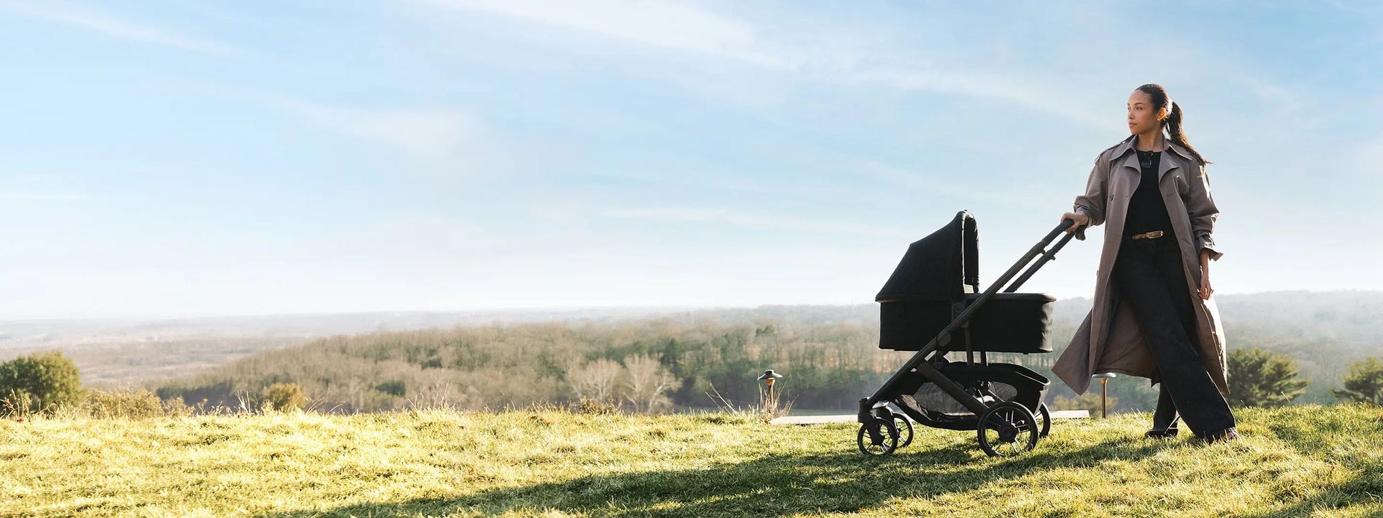 Mum in a trench coat walks a black pram with bassinet across a grassy hilltop under a wide blue sky