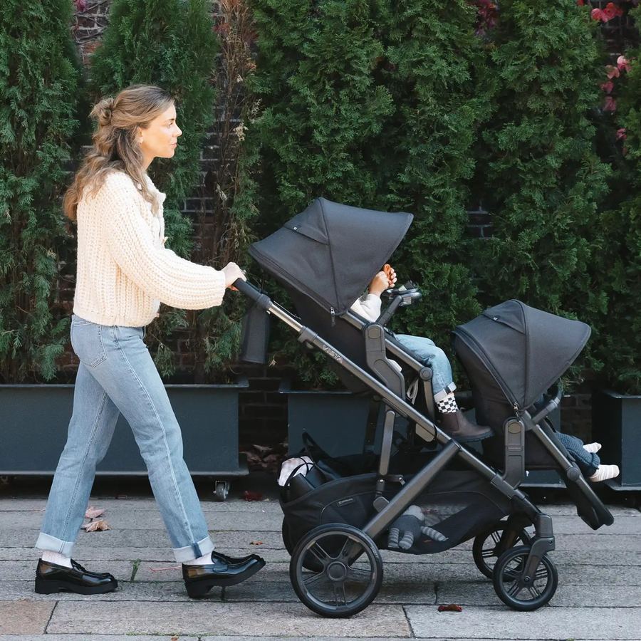 Side view of a mum walking a charcoal Vista V3 with two children in the toddler seat and RumbleSeat along a hedge-lined footpath